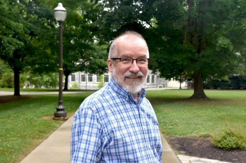 A man with balding salt and pepper hair and a beard with glasses and a plaid shirt stands on a sidewalk near some trees.