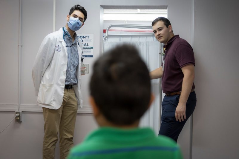 Two medical students standing at a vision screening poster, one poiting to an object on the poster. Back of young boy's head in foreground.