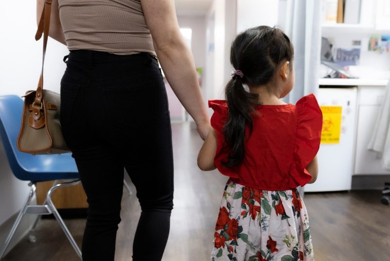 Back of little girl, holding her mother's hand walking down a clinic hallway
