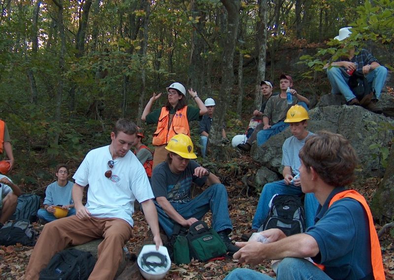 A group of people wearing safety gear sit in a forest. At center, a person is standing with arms raised.