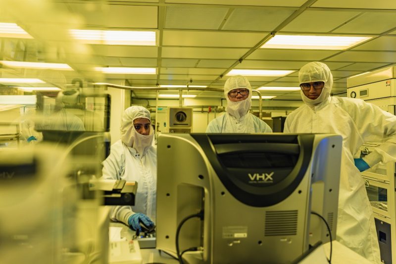 Student stands in clean room gown while holding open plastic case of semiconductor components
