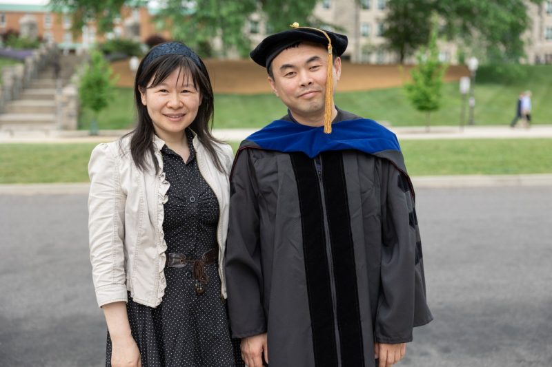 A female faculty member and male graduate student wearing Ph.D. robe stand outside with campus view in the background