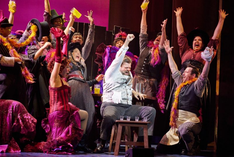 Alfred P. Doolittle sits with his fist triumphantly in the air, surrounded by many men and women dressed in saloon dresses, top hats, vests, and feather boas, who also have their hands in the air, faces bright with fun and triumph. This stage is scene is of the nationally touring company of "My Fair Lady." 