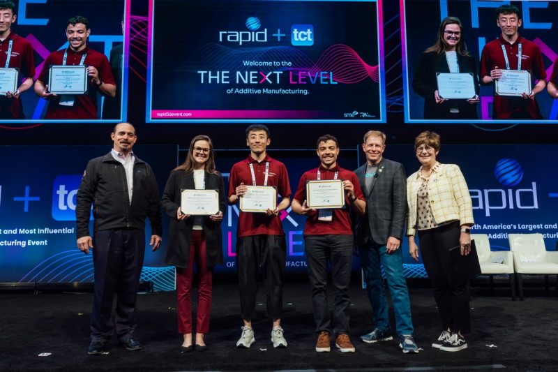 Six people stand together on a stage with a colorful, high tech digital background. The three center people are each holding certificates and smiling. 