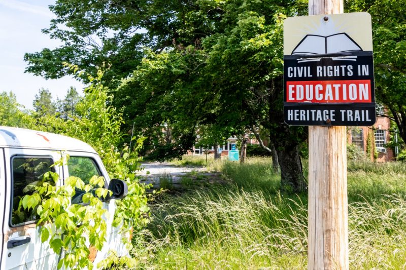 A sign that reads "Civil Rights in Education Heritage Trail" sits next to an abandoned security van with high grass, overgrown trees, and brick academic buildings in the background.