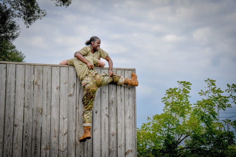 A woman new cadet hauls another new cadet over the team wall on the obstacle course.