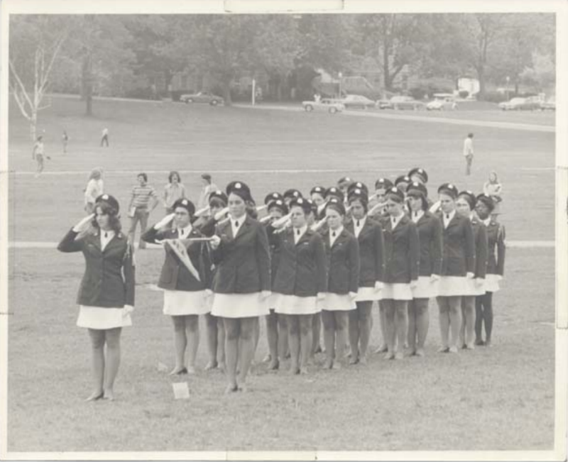 L Squadron stands on the Drillfield in formation. All the women are in dress uniform with white skirts and they are saluting.