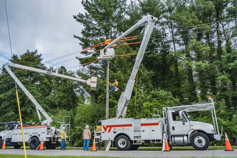 Two Virginia Tech Electric Service bucket trucks parked underneath power lines that are located next to many trees. Line workers work on the power lines and some are on the road observing the activities.