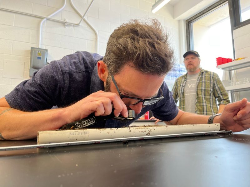 A man in na blue shirt closely examines some collected sediment using a magnifying glass.