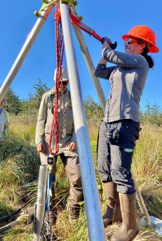 A woman, wearing a hard hat, use a three pronged , tall apparatus to insert deeply into the ground a metal tube for collecting core samples.