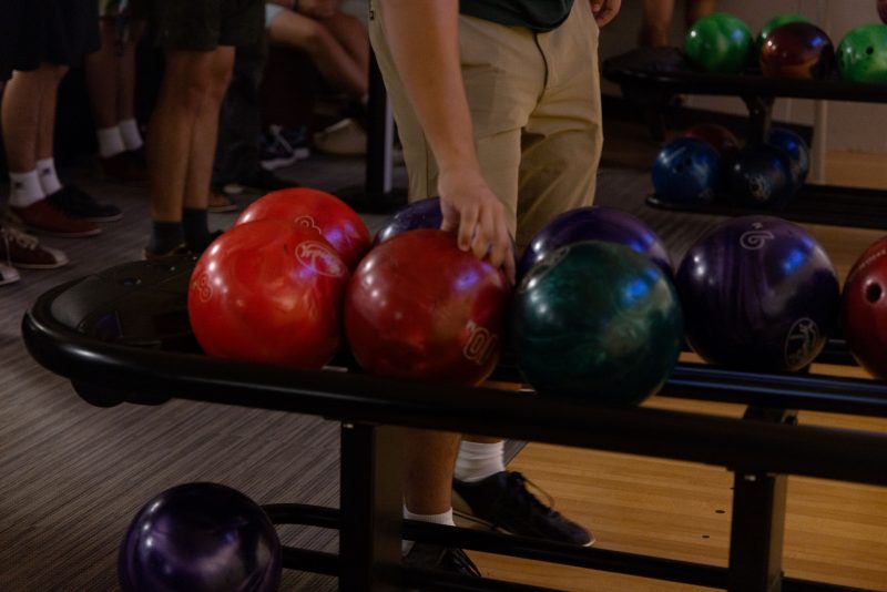 Several bowling balls in various colors are in the foreground. Behind them, you can see the arm of a student reaching down to retrieve one. The lower bodies of other students and more bowling balls can be seen in the background.