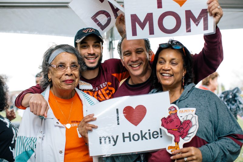 A family dressed in various colors and styles of Virginia Tech spiritwear poses for the camera and holds up signs with heart-shaped icons that say "I (heart) my Hokie!", "I (heart) DAD" and "I (heart) MOM"
