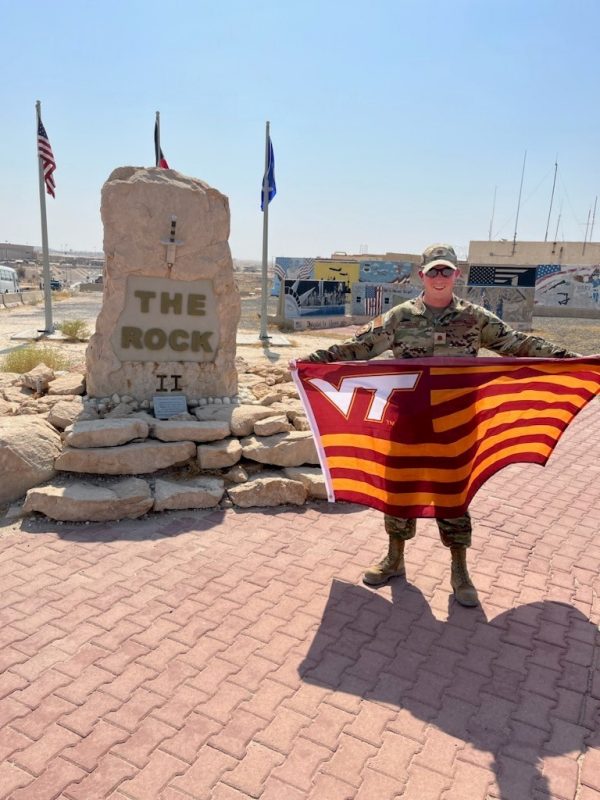 U.S. Air Force Major Kyle Hooper smiles while holding a Virginia Tech flag. He is wearing his camouflage uniform and sunglasses in a desert setting.