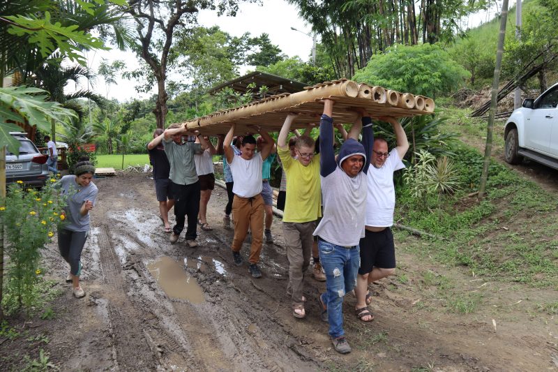 Students carrying large bamboo structure across muddy road.