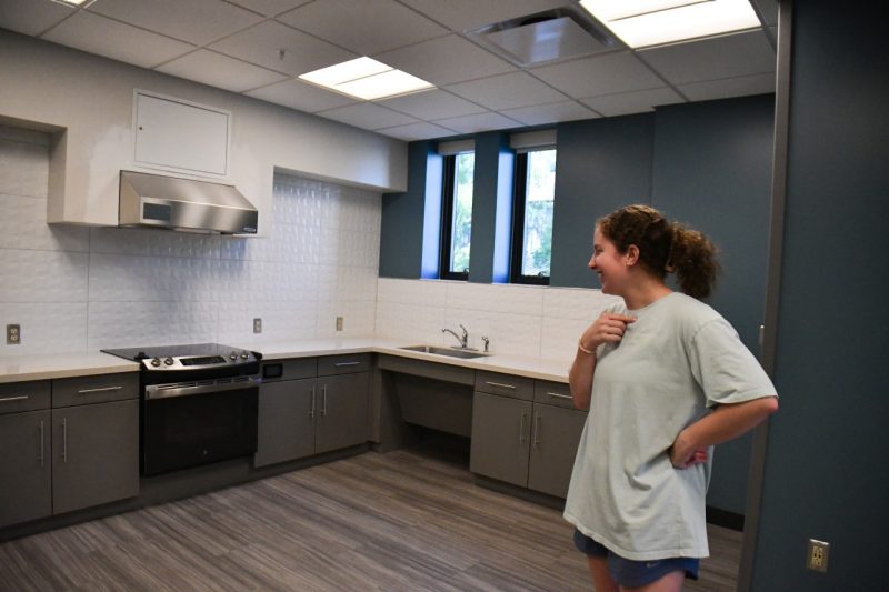 A female cadet in workout clothes smiles while looking at the kitchen, which is modern and clean. A white tile backsplash fills the space behind the stove.