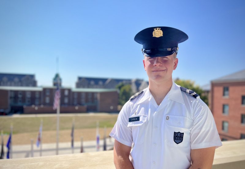 Cadet Carlin stands in his uniform on a balcony overlooking Upper Quad. Lane Hall is in the far background and the cadet is smiling.