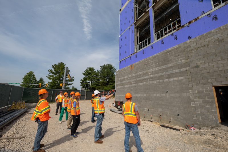 People wearing bright orange high visibility vests and hard hats gather outside on gravel. A building under construction is in front of the group. 