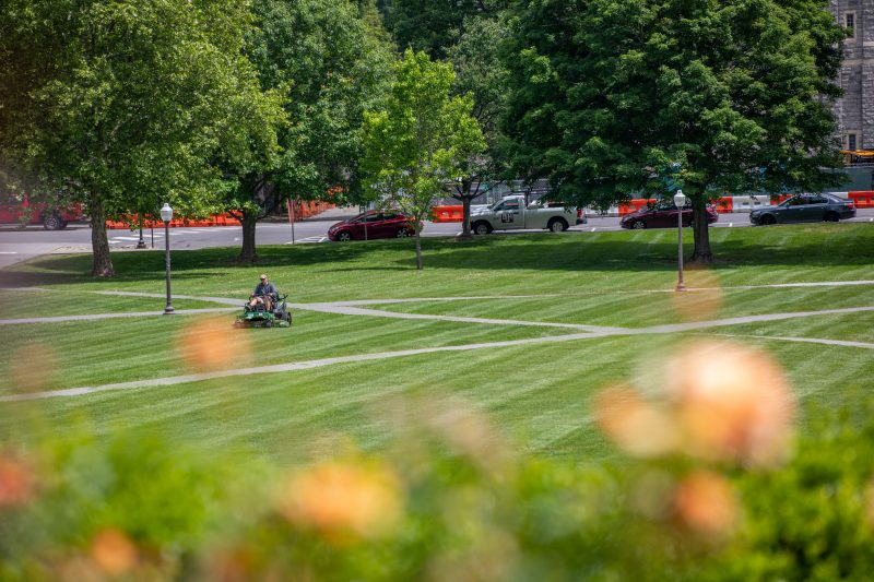 A person rides a lawn mower on the Drillfield on a sunny day. Blue skies with fluffy white clouds are above. 