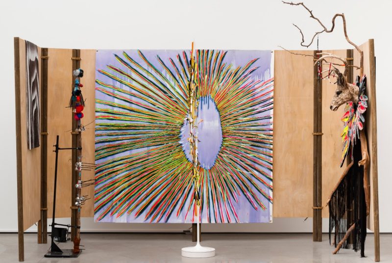 An installation view of Laurie Steelink's "Gathering Power," 2022. A three-paneled wood structure on which hangs two paintings. The painting in the center is a multicolored starburst or dream catcher pattern. Sculptural elements at left and in the center are reminiscent of totem poles. At right, a deer head and a willowy tree branch hang on a panel.