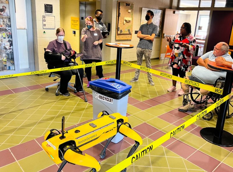 Consultants and researchers gathered around an indoor caution-taped area, watching a yellow robot dog and trashcan obstacle.