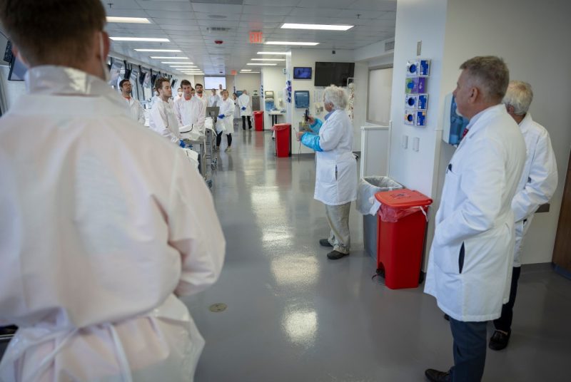 Long shot in lab setting.; students, in white lab attire on the left, listening to instructors on the right