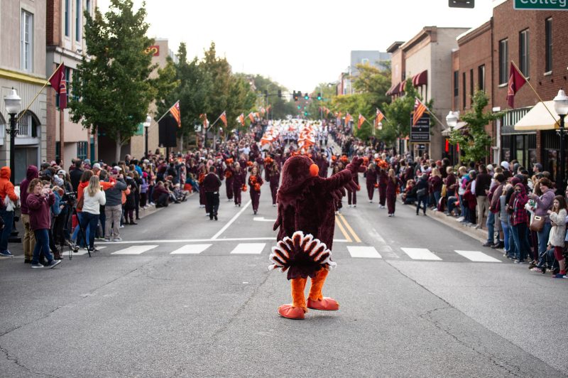 The HokieBird, facing away from the camera, gestures towards a large crowd of people on either side of a street in Blacksburg, mostly dressed in Virginia Tech colored spiritwear. Behind him are marching cheerleaders wearing maroon warm-up suits and waving pom-poms. In the distant background is a marching band dressed in white and maroon uniforms and holding Virginia Tech-colored flags.