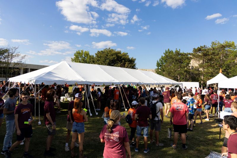 A group of students in Hokie spiritwear form a line to enter a large white canopy tent behind them. Behind the tent is a blue sky with scattered clouds, green trees and the tops of Hokie stone buildings.