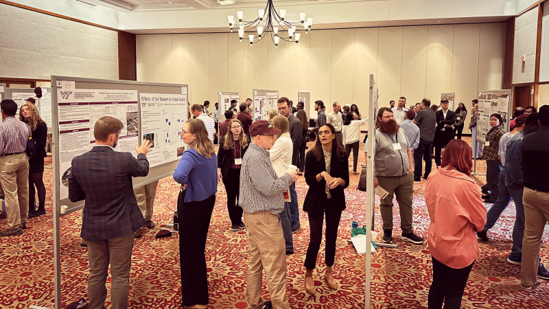 Students present their research posters to a panel of judges in a large room with maroon patterened carpet.
