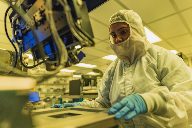 A student wearing a protective suit uses a machine to fabricate a semiconductor in a lab.