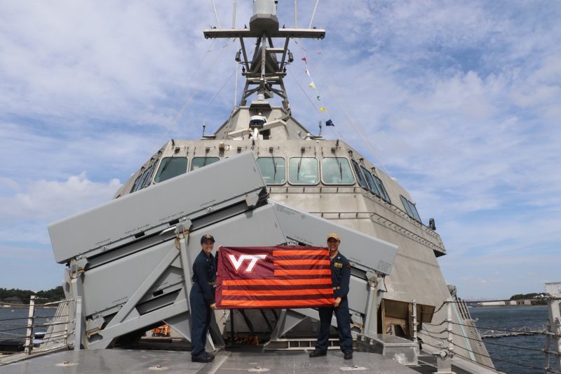 Miller and Nguyen stand holding a striped VT flag on a Navy ship. Both are smiling and wearing military uniforms.