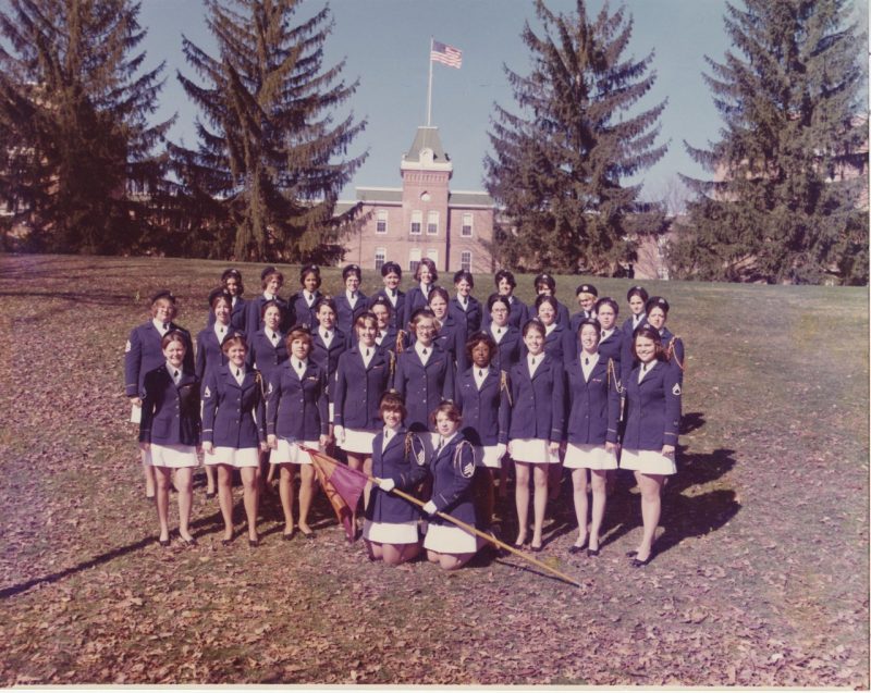 Women cadets in white skirts and dress blouses stand on the edge of Upper Quad below Lane Hall.