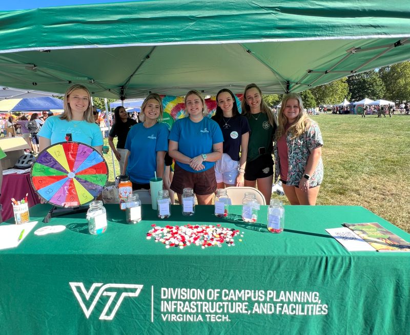 Many students smile in a group behind a table underneath a popup tent on Virginia Tech's Drillfield. There are interactive games on the table.