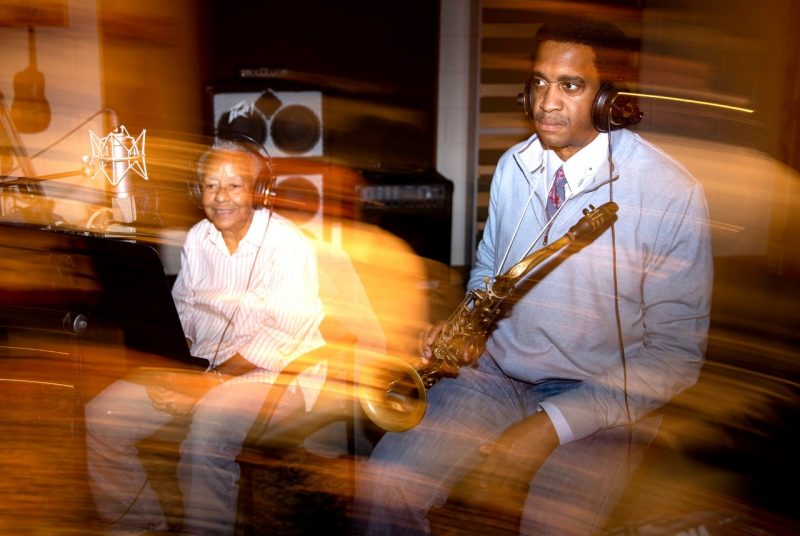At left, Nikki Giovanni, an older Black woman with short, grey, natural hair, and at right, Javon Jackson, a Black man with a short Afro. They sit in a recording studio, both wearing headphones, and he holds his saxophone. Blurred yellow lights reflect in front of the pair.