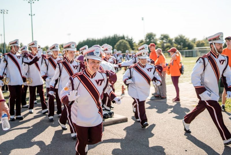 A large group of band members walk down a concrete path. They are all dressed in Marching Virginians uniforms - white tops with maroon sashes accentuated with a maroon VT logo on one side, maroon pants, and white and maroon hats.