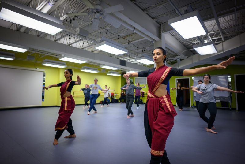 Two dancers from Chitrasena Dance Company lead a dance workshop. The two women both have brown skin and dark hair and both are wearing a burgundy sash wrapped around their shoulders and wait, down to their legs over black cropped shirts and black pants. They each strike poses with their hands stretched out to their sides. A group of students mimic their pose behind them.