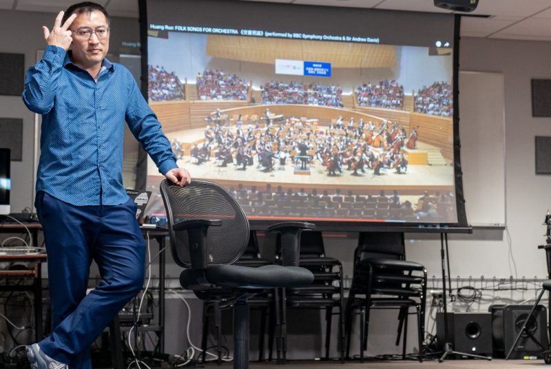 Composer Huang Ruo stands in front of a projector screen showing an image of an orchestra. He wears glasses and a blue button-up shirt and dark blue pants, leaning against an office chair.