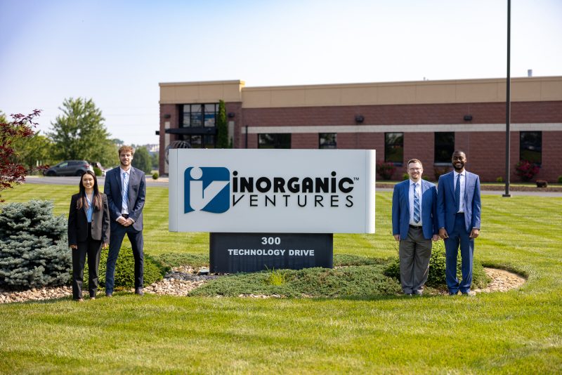 Four students dressed up in professional attire stand on either side of a large sign reading "Inorganic Ventures, 300 Technology Drive."