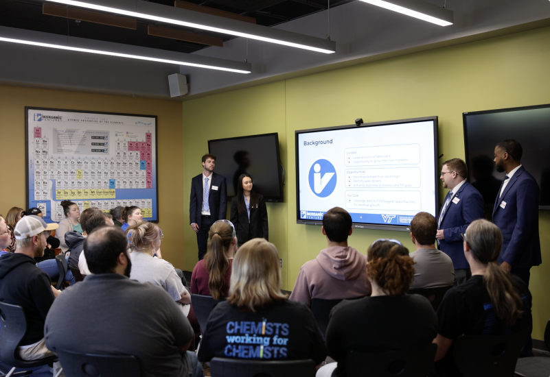 Four students in professional attire stand in front of a large group of Inorganic Ventures employees who are seated and watching the presentation.