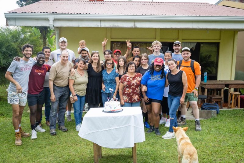 Students and Costa Rican community members pose for a photo outside a yellow home with a construction-themed cake. 