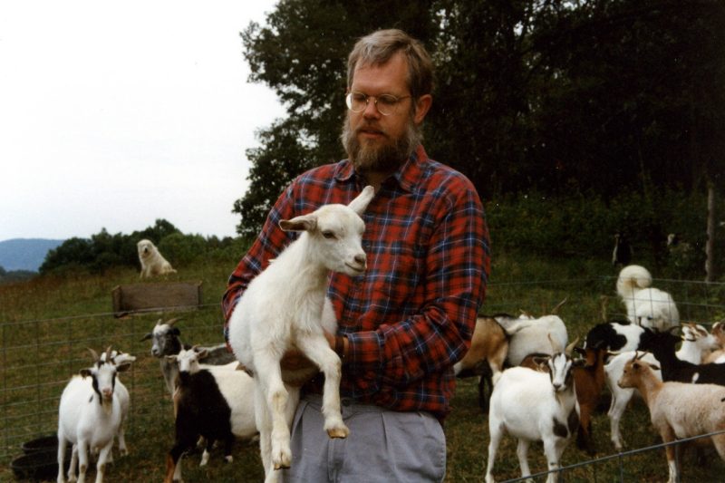 Person in a flannel shirt, holding a goat and goats standing behind them.