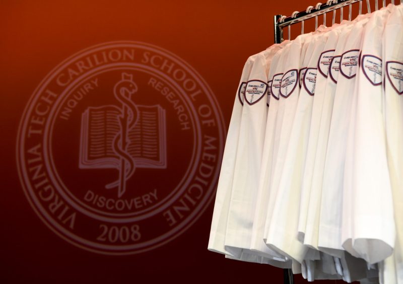 The Virginia Tech Carilion School of Medicine seal projected onto a wall in orange behind a rack of white coats.
