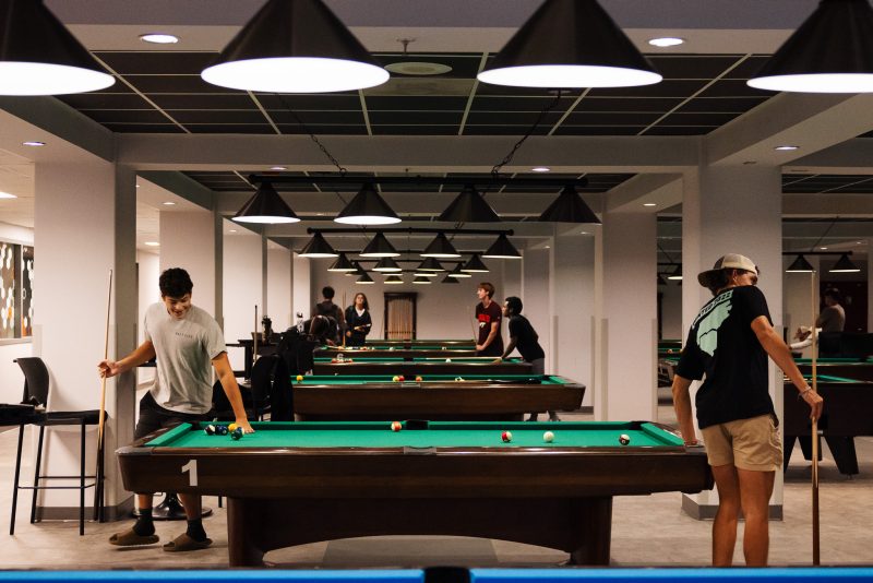 The camera looks down a long row of pool tables positioned between columns. Several students in casual wear are actively playing pool on them.