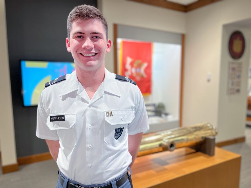 Cadet Hutchison stands in his uniform smiling. Museum items are in the background, including a red flag in a display case and the brass barrel of the original Skipper canon.  