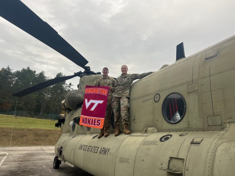 Young and Wolfe stand on the side of a military helicopter that is sitting on the ground. They are in camouflage uniforms holding a VT flag and both are smiling.