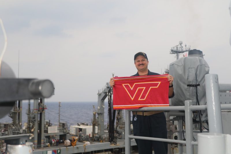 Taylor smiles while holding a Virginia Tech flag. He is wearing his blue Navy uniform and standing on a ship at sea with another ship in the background.