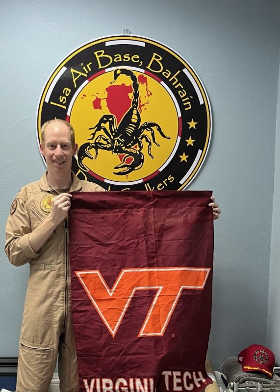 Frost stands in front of a blue wall with a sign saying Isa Air Base, Bahrain. He is holding a Virginia Tech flag while he smiles in a beige military flight suit. 