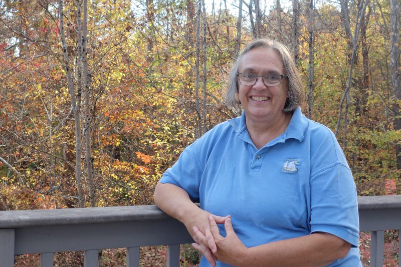 Woman with short hair and a blue polo shirt stands smiling next to a deck railing with trees in background