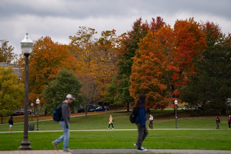 Bright orange, red, and green leaves cover the trees surrounding the Drillfield. In the foreground, students walk across the Drillfield out of focus/ 