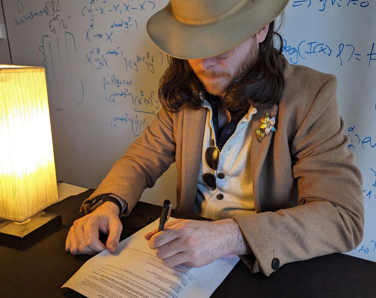 
A man with a hat pulled low over his eyes signs a paper at a table. The whiteboard in the background is covered in equations.
By adding a simple algebraic twist into the equation, mathematics' Jason LeGrow ventures into deeper territories of number theory to protect digital interactions from a large-scale quantum attack.
