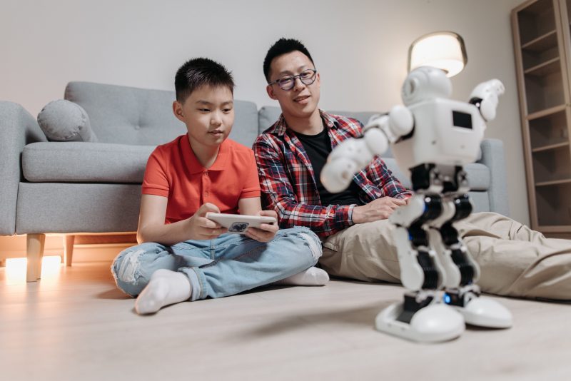 A man watches a boy play with a toy robot. Image courtesy Pexels.